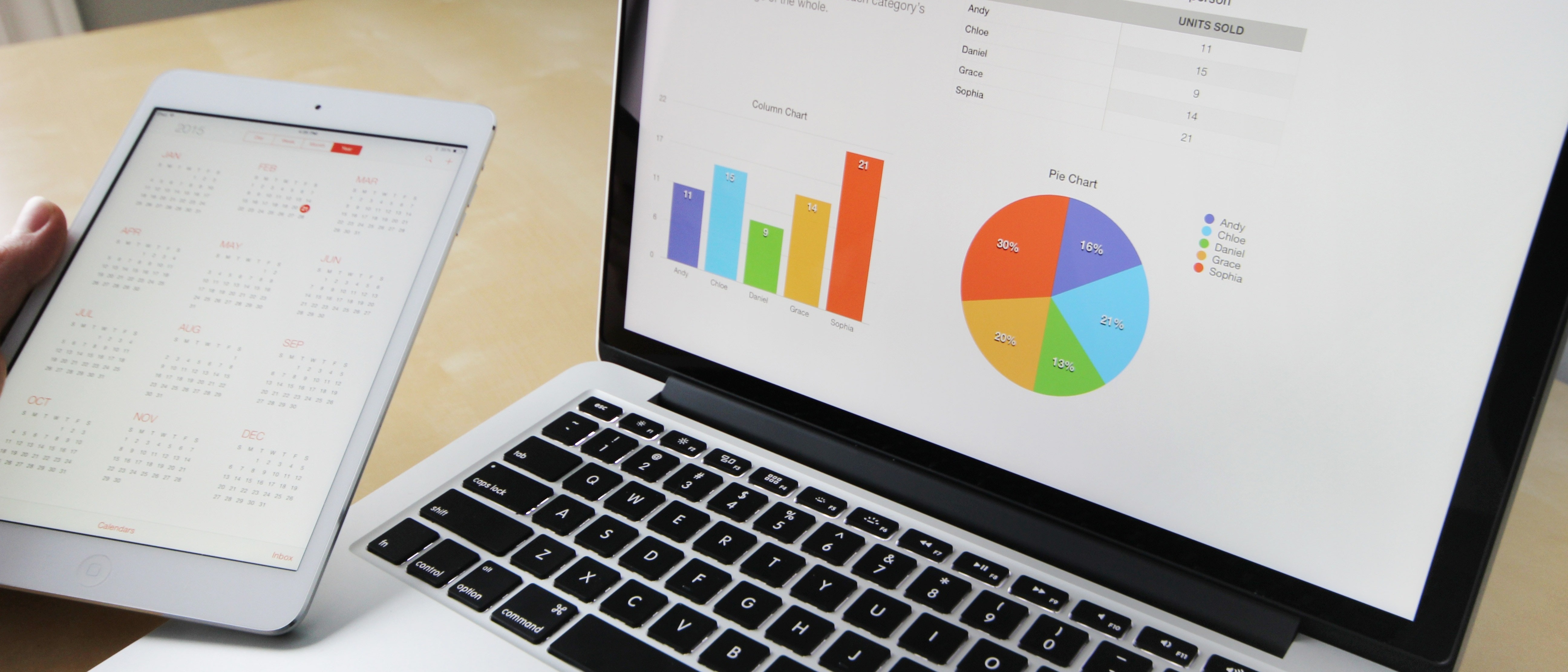 A person holds a tablet showing a calendar beside a laptop displaying colorful bar and pie charts